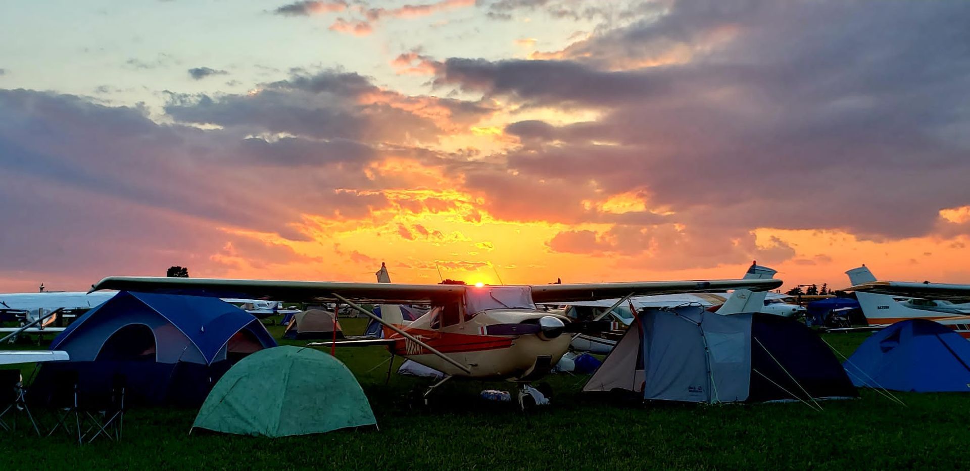 Tents and parked cars silhouetted at sunset in an open field with colorful clouds.
