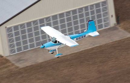 A smiling person wearing a blue shirt stands in front of a red airplane inside a hangar, with event title text overlaid.