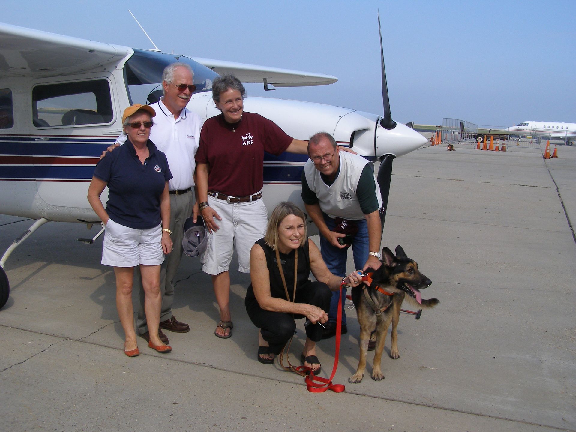 A group of five people and a German Shepherd stand on an airport tarmac next to a small white and blue airplane.