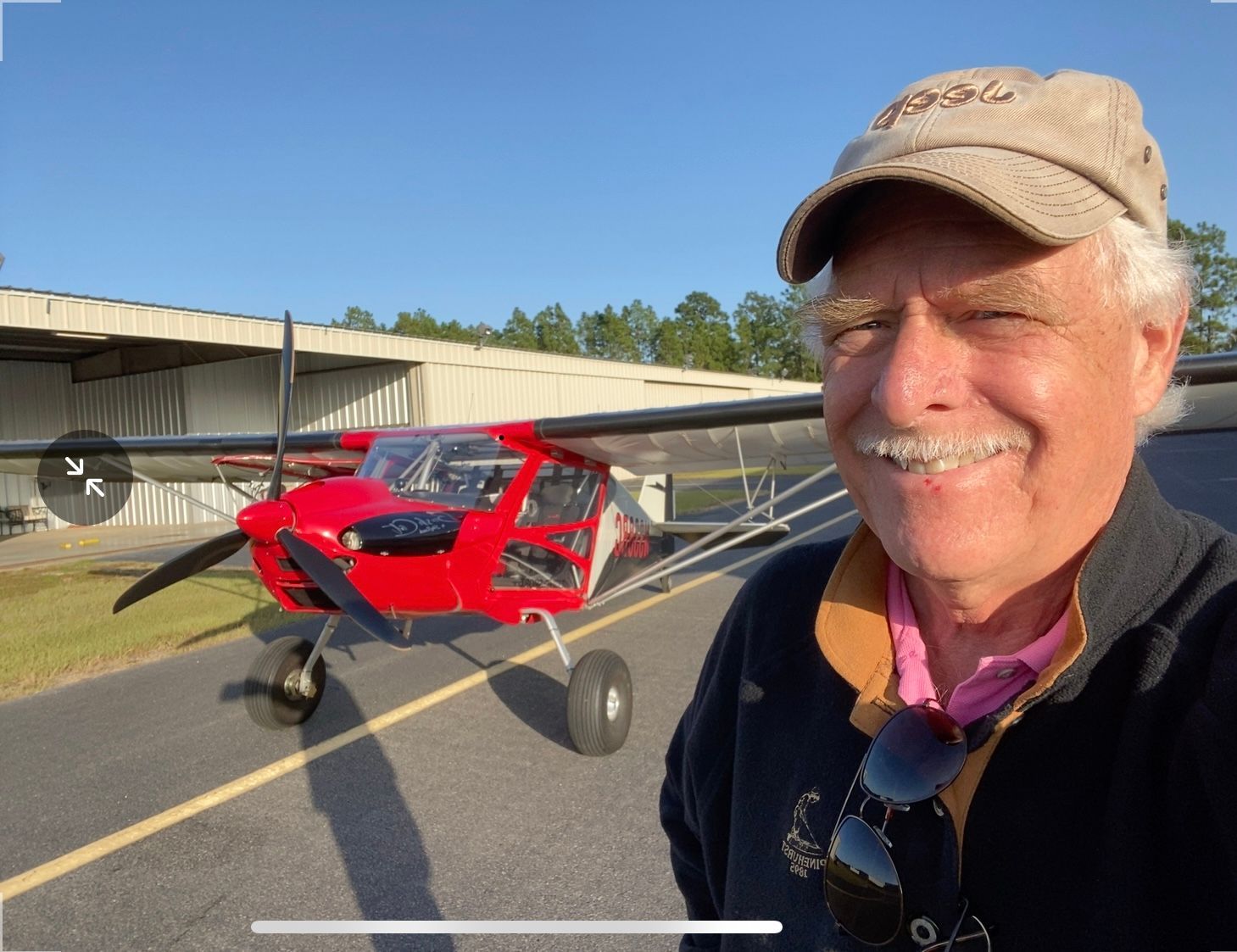Smiling man in a cap posing beside a red light airplane on a runway, with blue sky behind.
