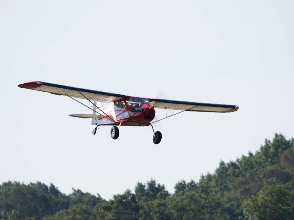 A red and white high-wing light aircraft flying above a treeline against a pale sky.