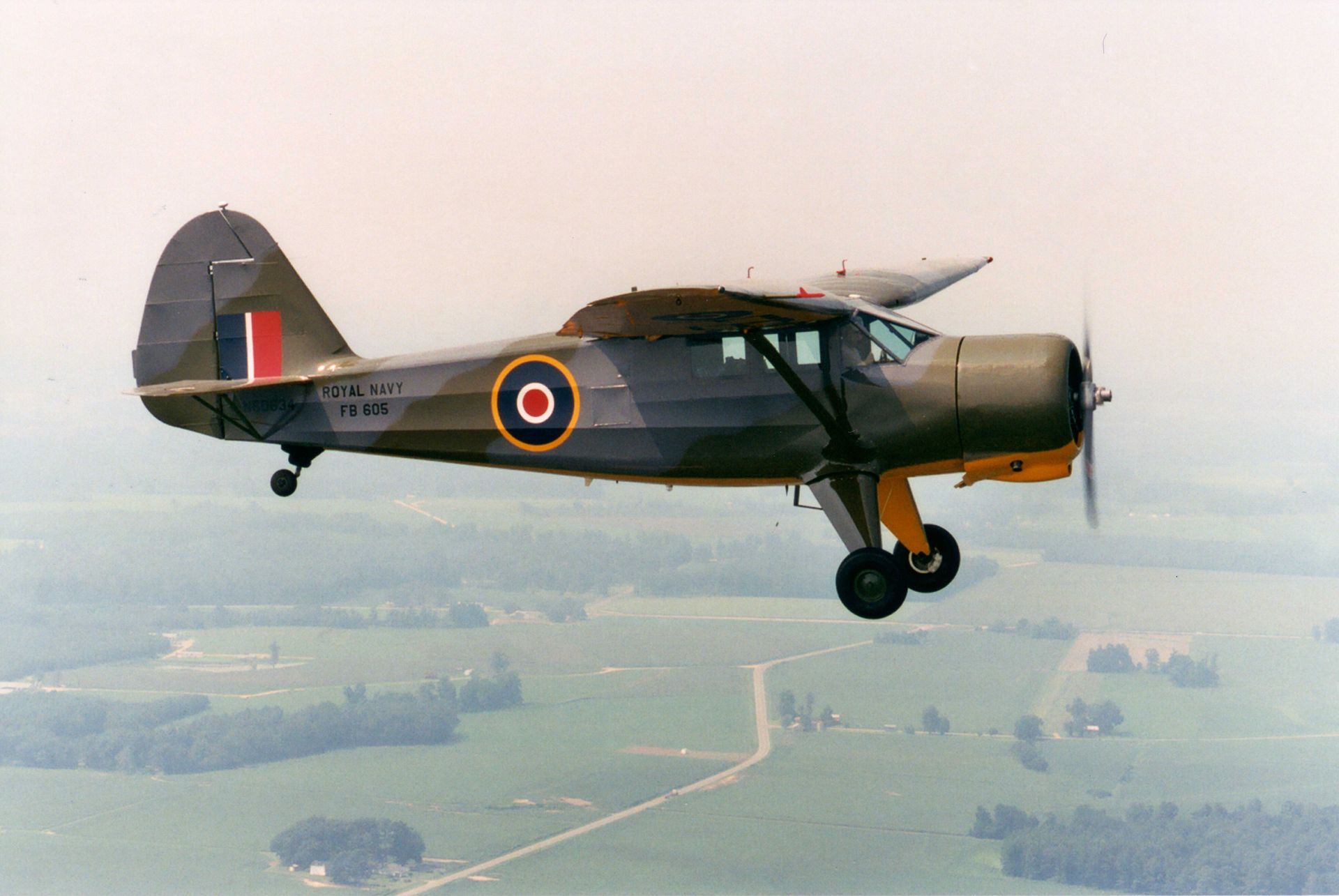 Vintage military aircraft flying over countryside, painted dark green with RAF roundel markings.