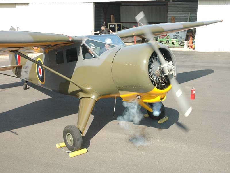 A vintage military aircraft with a green camouflage paint scheme, parked on an apron with smoke visible beneath the engine.