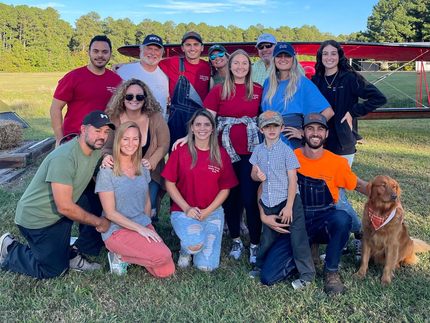 A large group of people posing in an outdoor field, some smiling, in front of a parked aircraft with a dog sitting nearby.