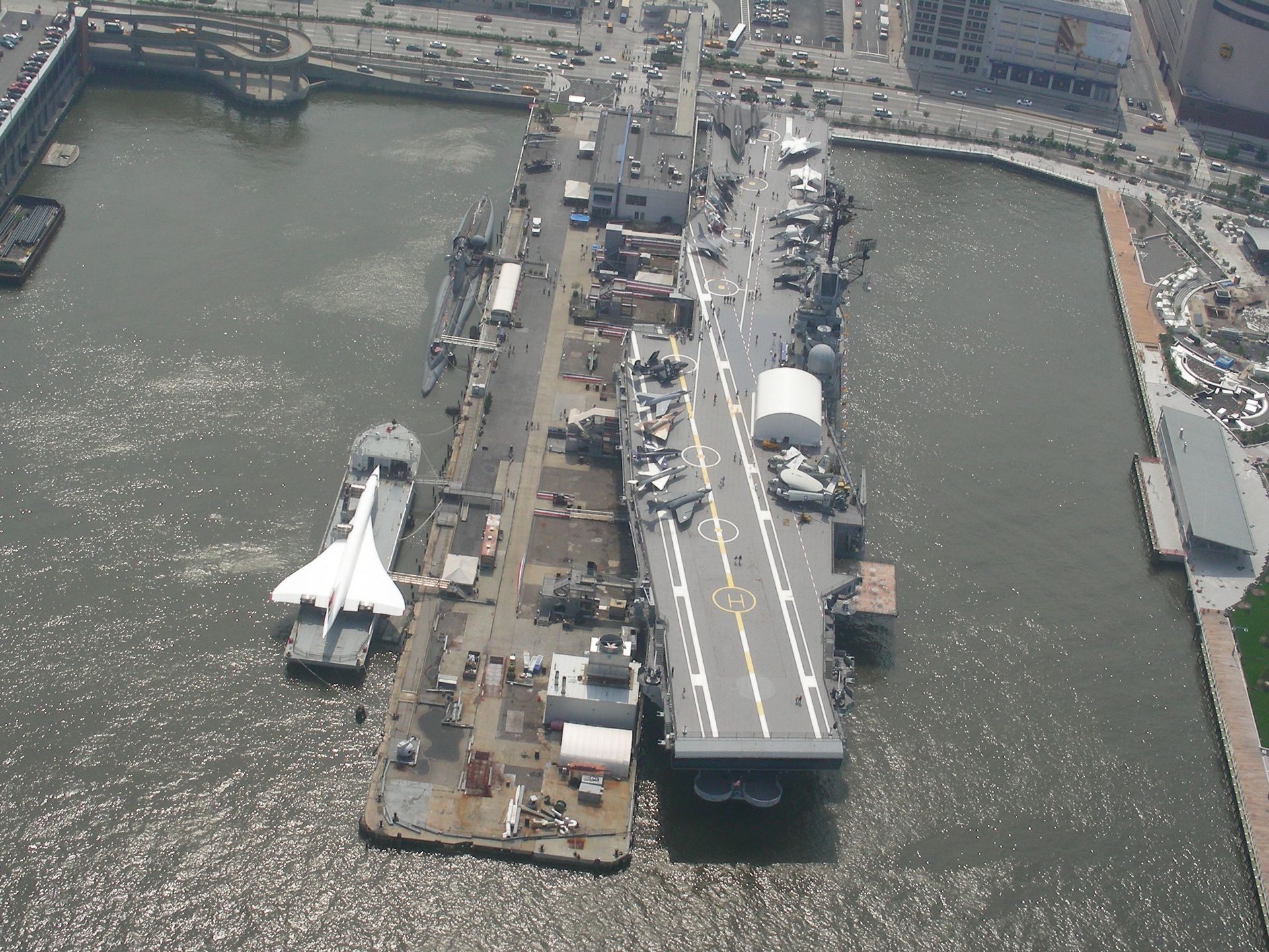 Aerial view of a docked gray warship in a harbor beside a waterfront pier and smaller boats.