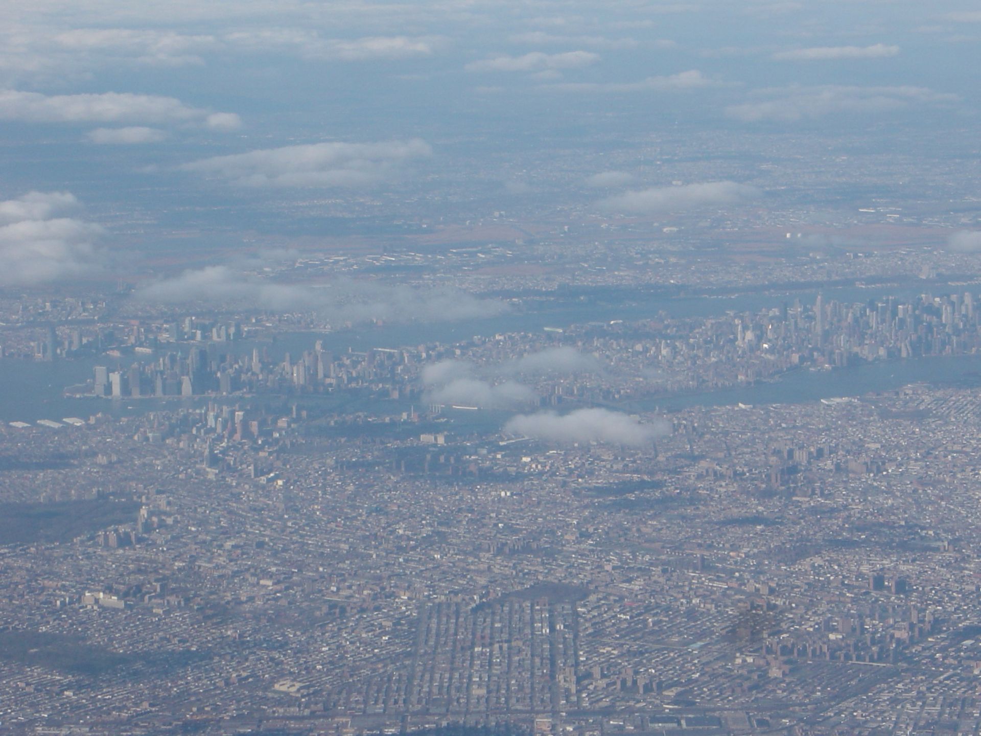 Aerial view of a sprawling city under a hazy sky, with clouds and distant water visible.