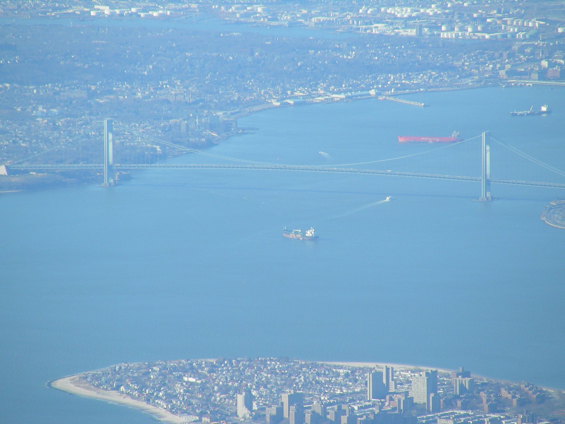 Aerial view of a suspension bridge over blue water with a city shoreline in the distance.