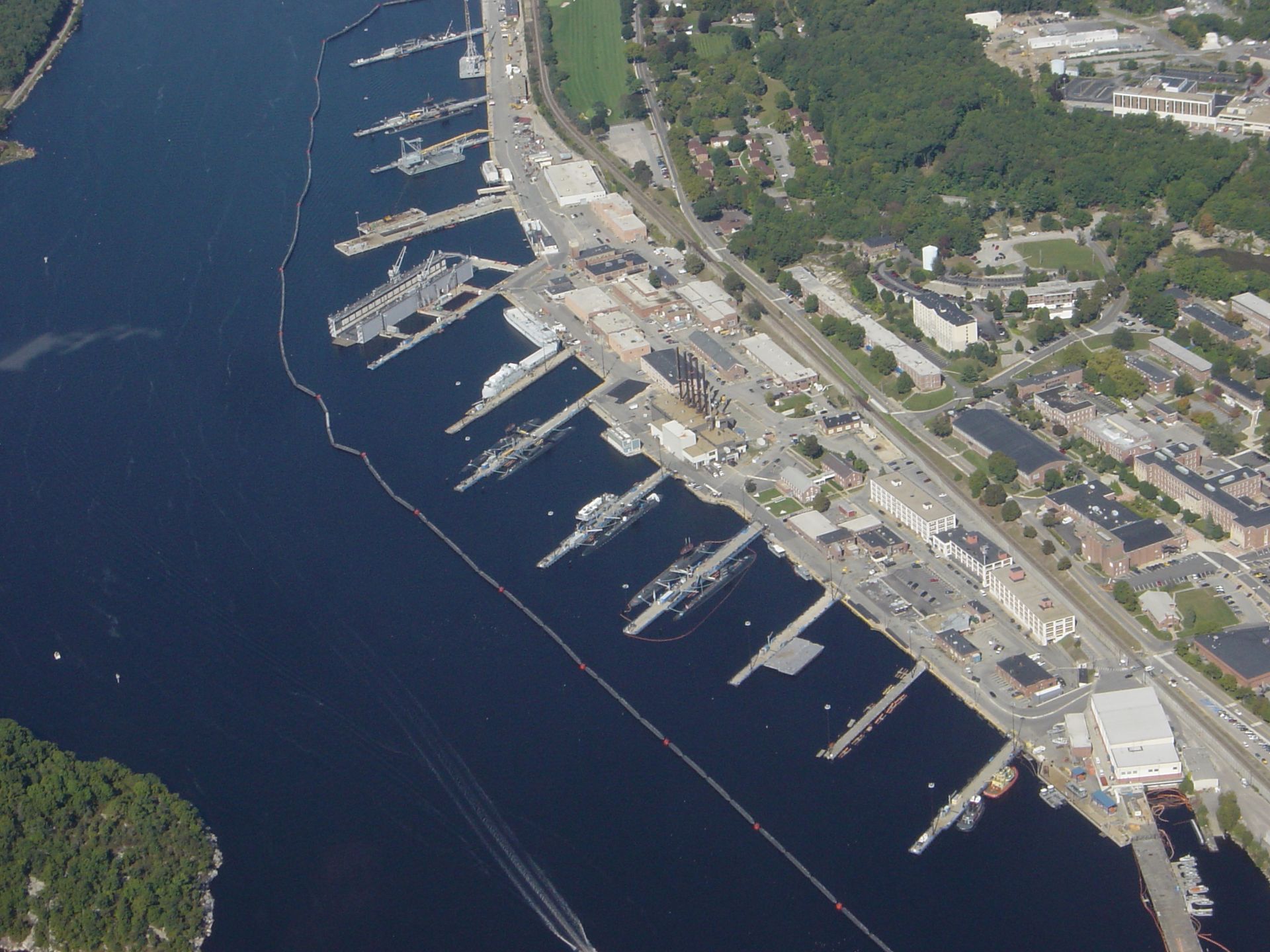 Aerial view of a marina with docks, boats, a waterfront road, and wooded shoreline