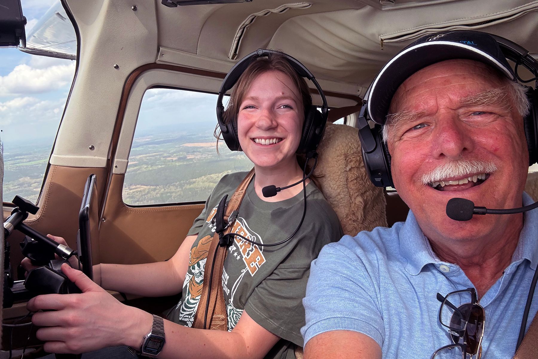 A smiling instructor and student sit in the cockpit of a small airplane, wearing headsets while in flight.