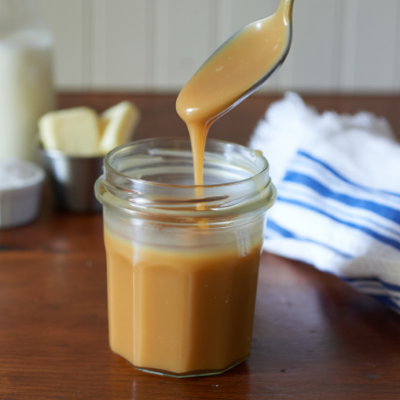 Spoon lifting caramel sauce from a glass jar on a wooden table