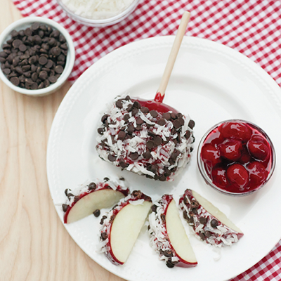 Chocolate-dipped apple slices and candy apple on a white plate with cherries and chocolate chips in the background