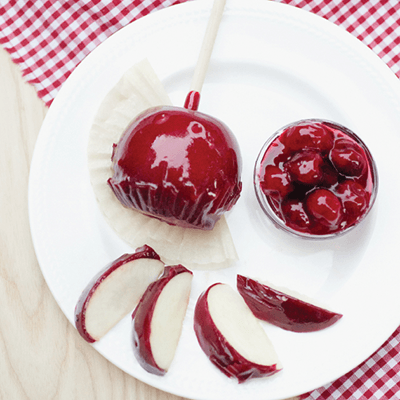 Candied red apple on a stick with sliced apple and cherry garnish on a white plate