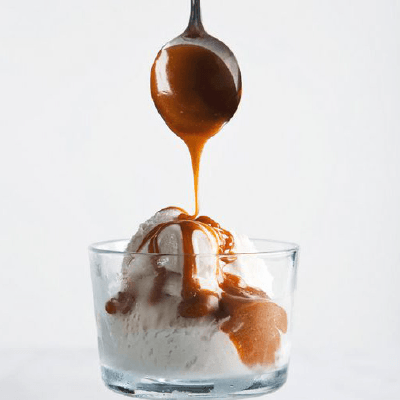 Vanilla ice cream in a glass cup being topped with caramel sauce from a spoon