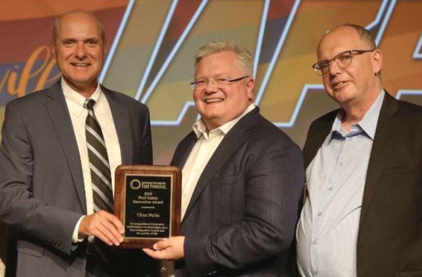 Three men in suits pose with a plaque award onstage against a geometric backdrop.