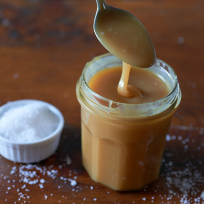 Spoon lifting creamy caramel from a glass jar beside coarse salt on a wooden table