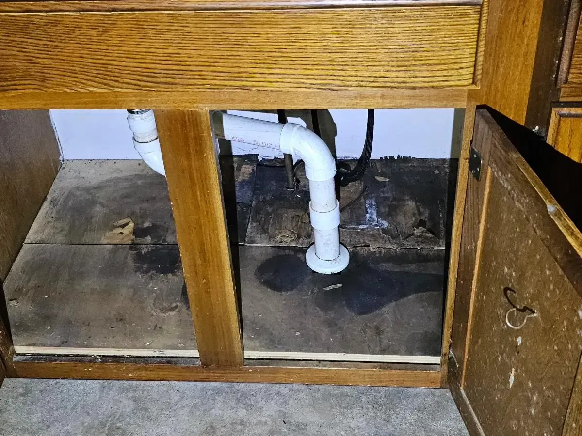 Under-sink plumbing with white PVC pipes beneath a wooden cabinet and open access panel.