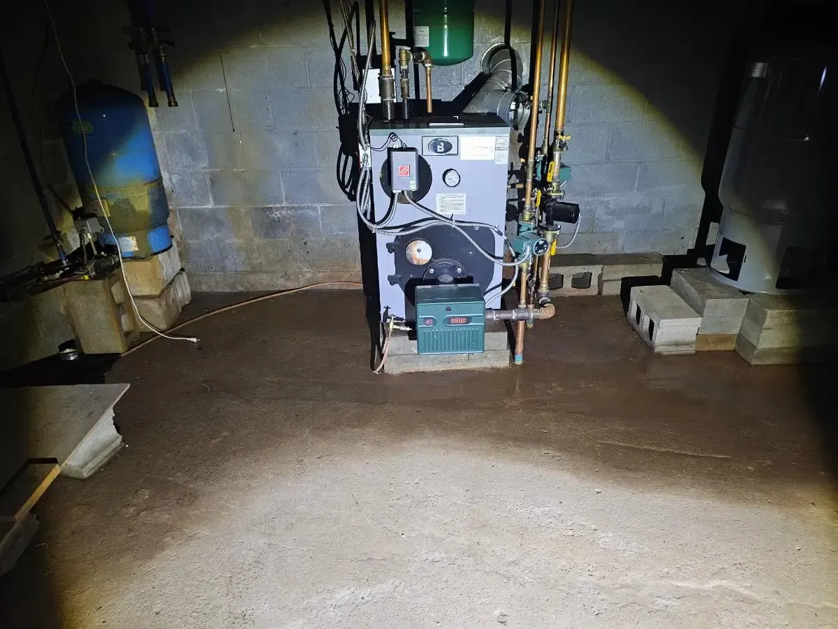 Dark basement with water on concrete floor; a heating system is centered with pipes and a blue water tank.