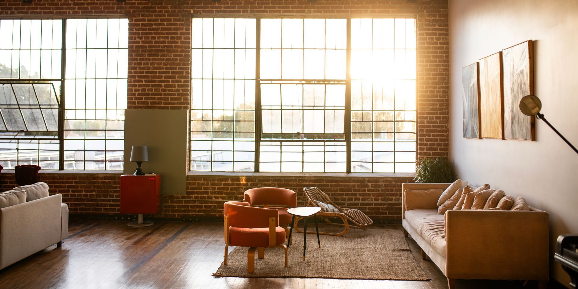 Sunlit loft living room with large windows, brick walls, tan sofa, orange chairs, and a red cabinet