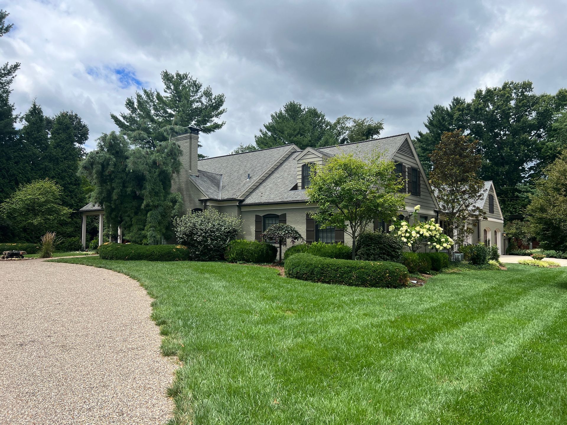 A house with a gravel driveway, green lawn, and trees on a cloudy day.
