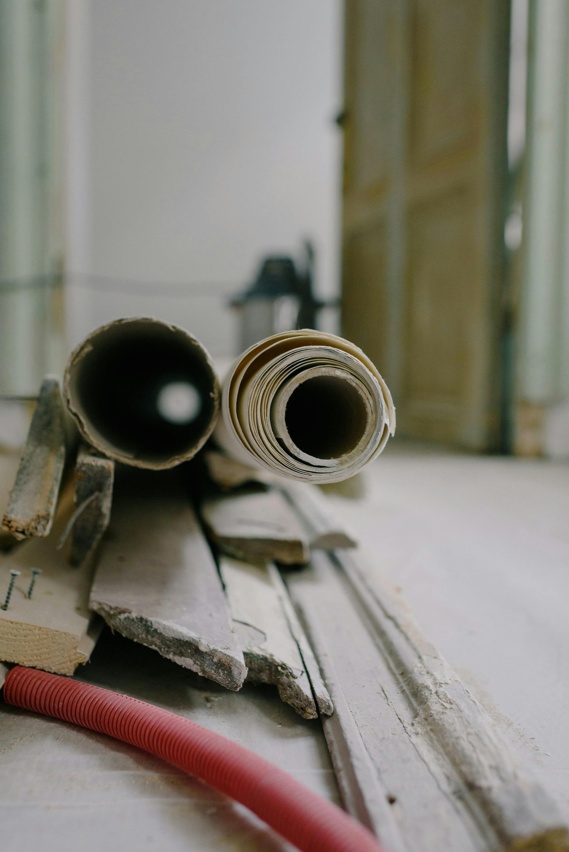 Rolled blueprints and wood scraps on floor, red hose in foreground, room under renovation.