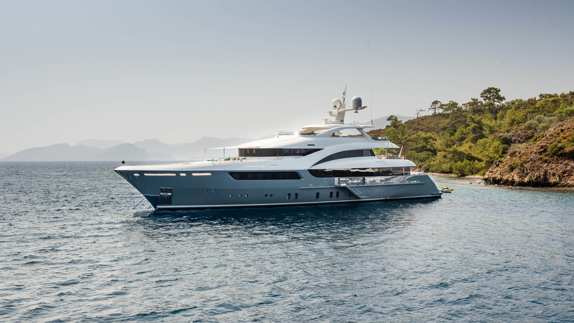 Luxury yacht on calm blue water near a tree-covered shoreline.