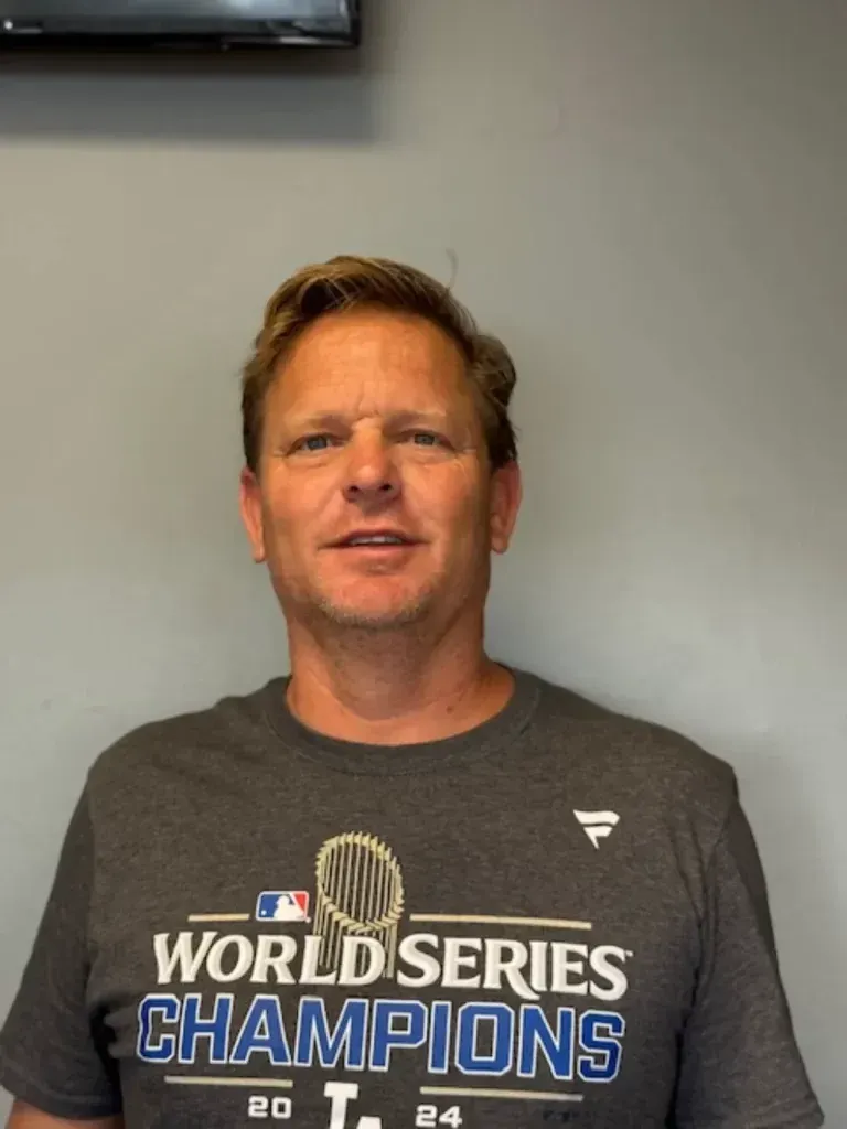 A man with blonde hair smiles in a grey Los Angeles Dodgers 2024 World Series Champions t-shirt against a grey wall.