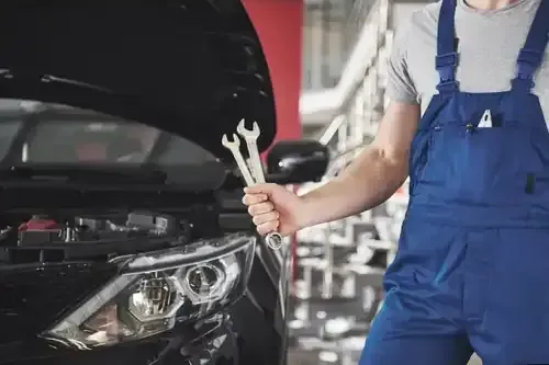 Mechanic in blue overalls holding a wrench beside a car with its hood open