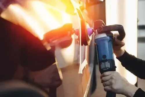 Person using a blue electric polisher on a car door, with bright reflections and sparks nearby