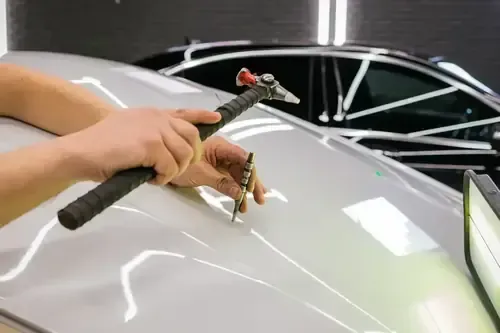 Hands using a paintless dent repair tool on a white car hood in a garage