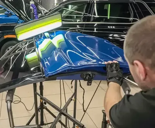 Person applying vinyl wrap to a blue car hood in a workshop