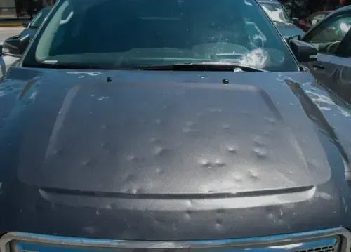 Gray car hood covered in numerous hail dents in a parking lot