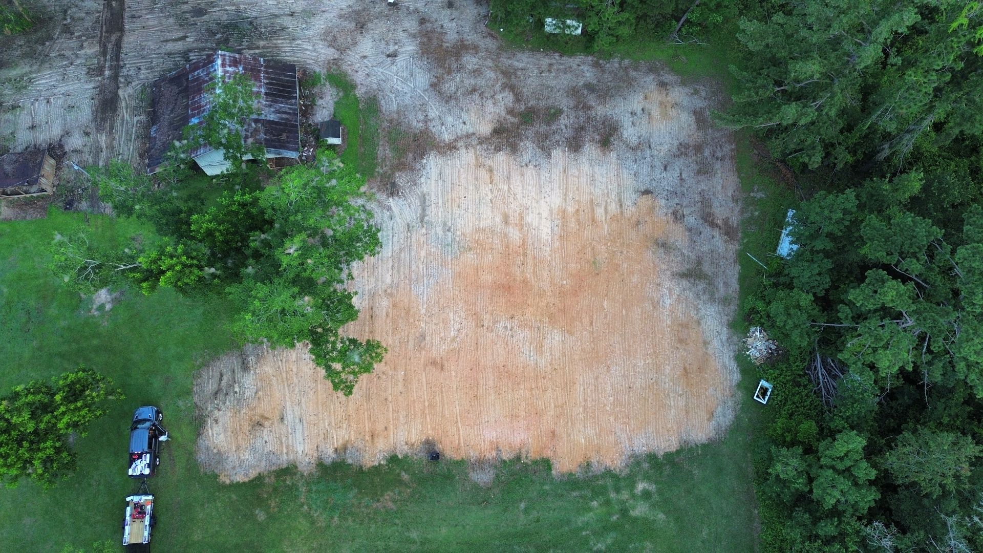Aerial view of a large tan dirt field surrounded by green trees and grass, with a few small people and vehicles at the edges