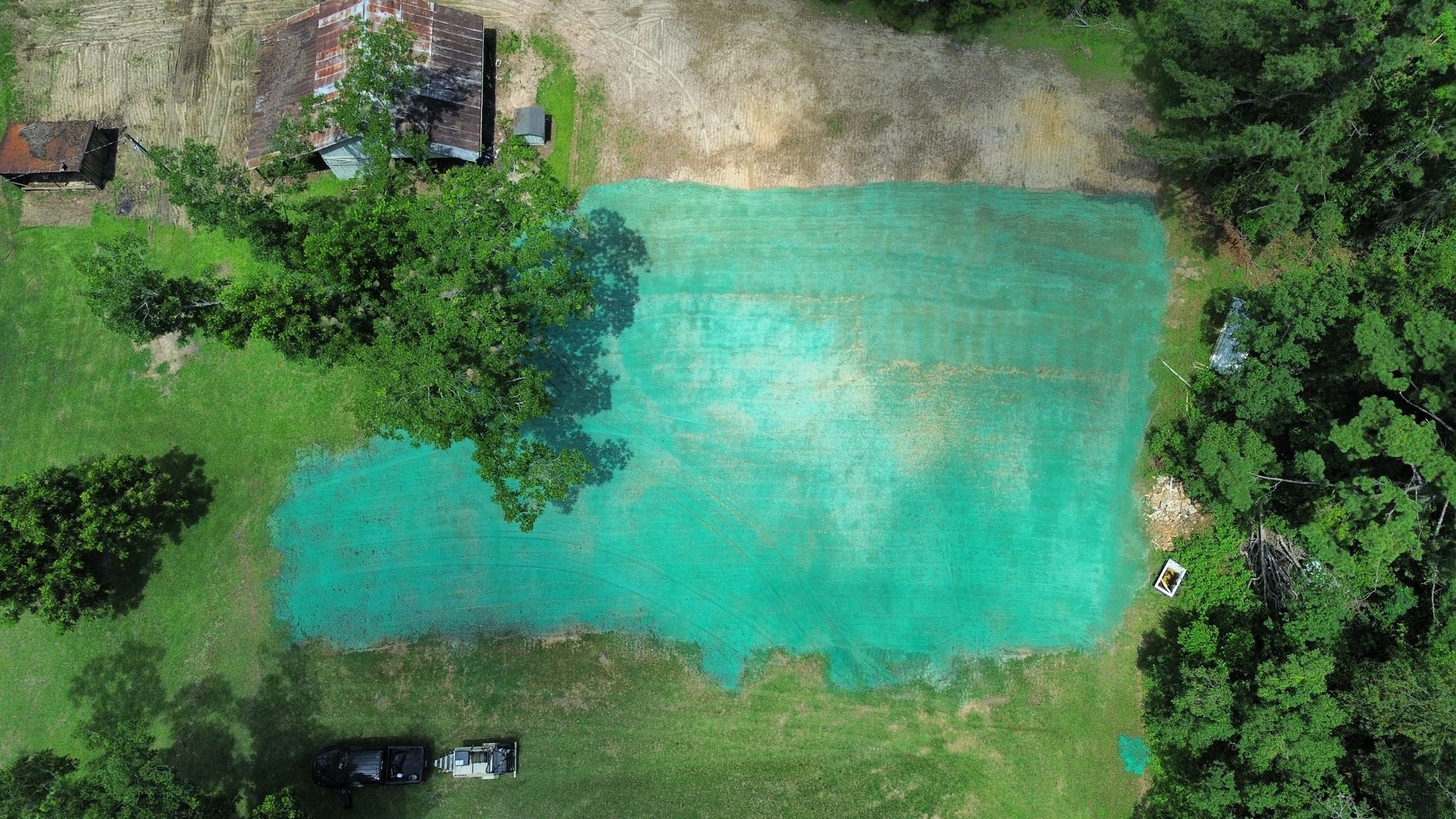 Aerial view of a turquoise swimming pool in a grassy yard, surrounded by trees and a few parked vehicles.