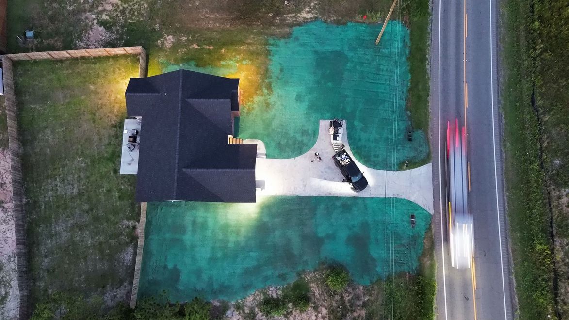 Aerial view of a house with a driveway, green yard, and a road along the right side.