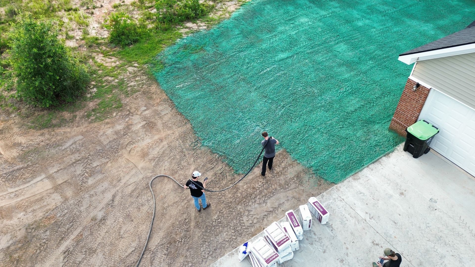 Overhead view of two people on a dirt yard beside a building, with green netting and construction materials nearby