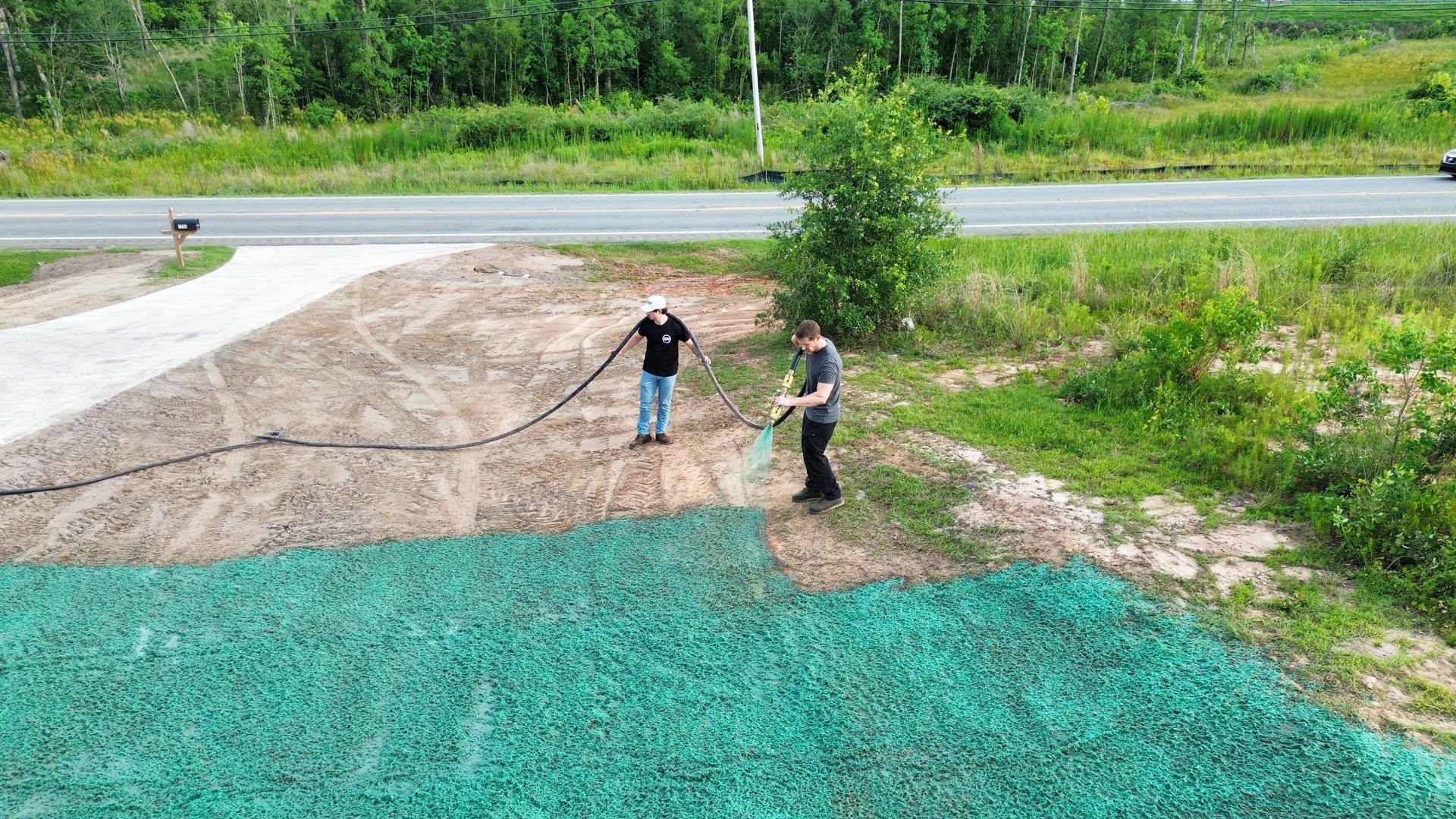Two people stand on a gravel pile near a road, with green mesh in the foreground and trees behind.
