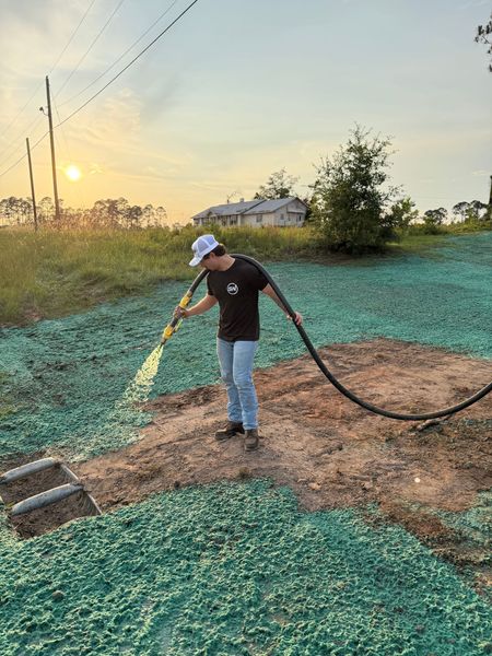 Person watering a garden bed at sunset with a hose, surrounded by green plants and dry soil.