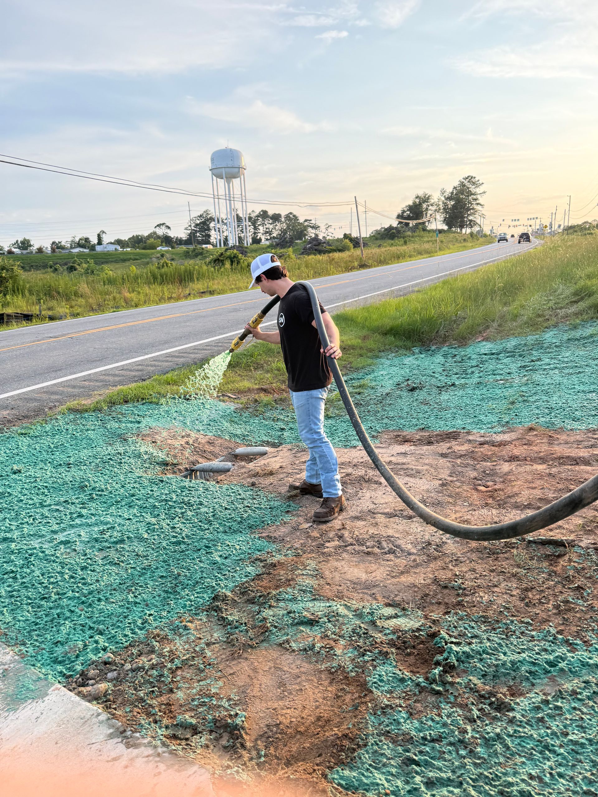 Worker spraying green material on roadside ground beside a water tower at sunset