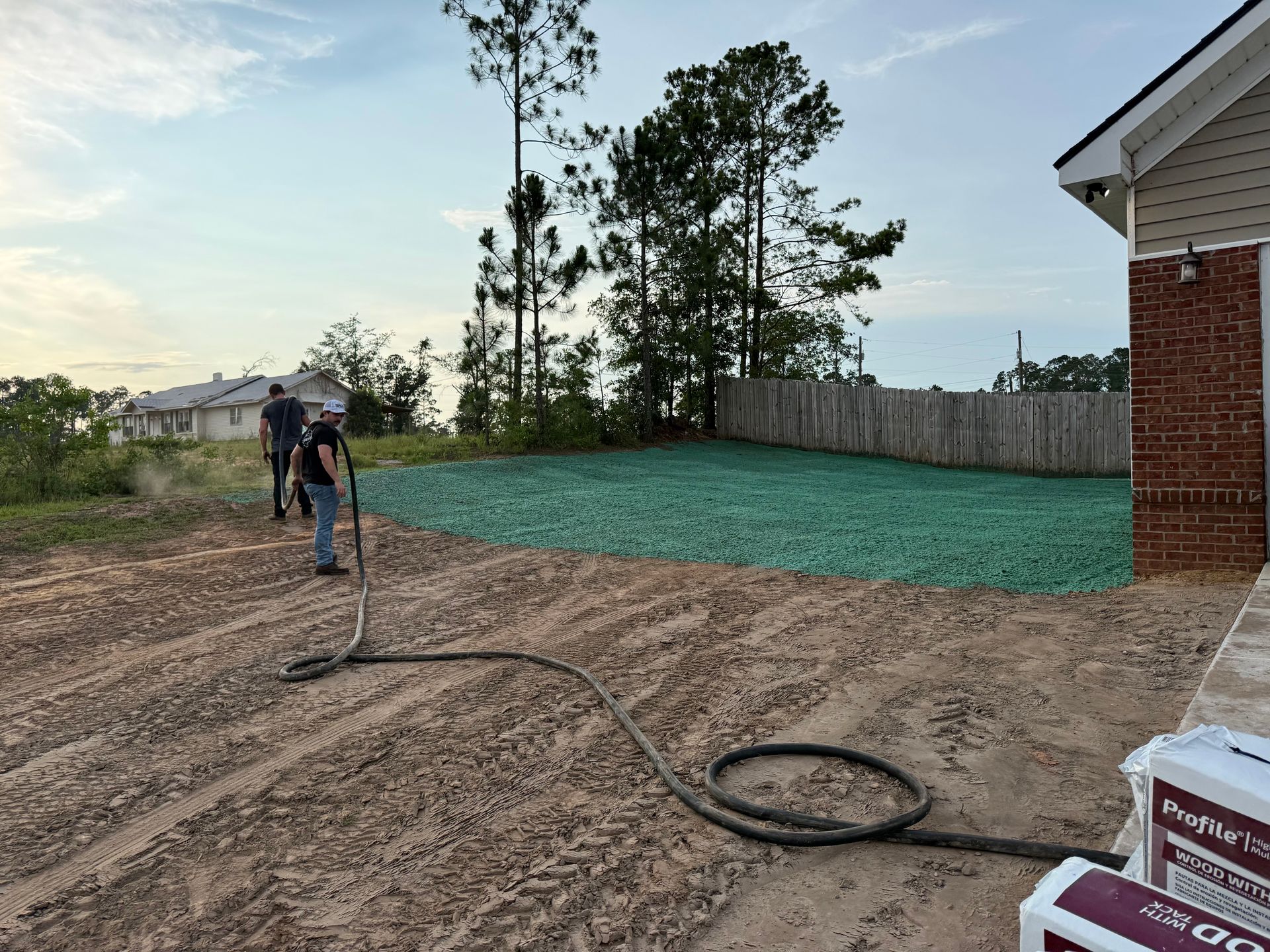 Worker spraying dirt in a backyard construction site beside a house and fence at dusk