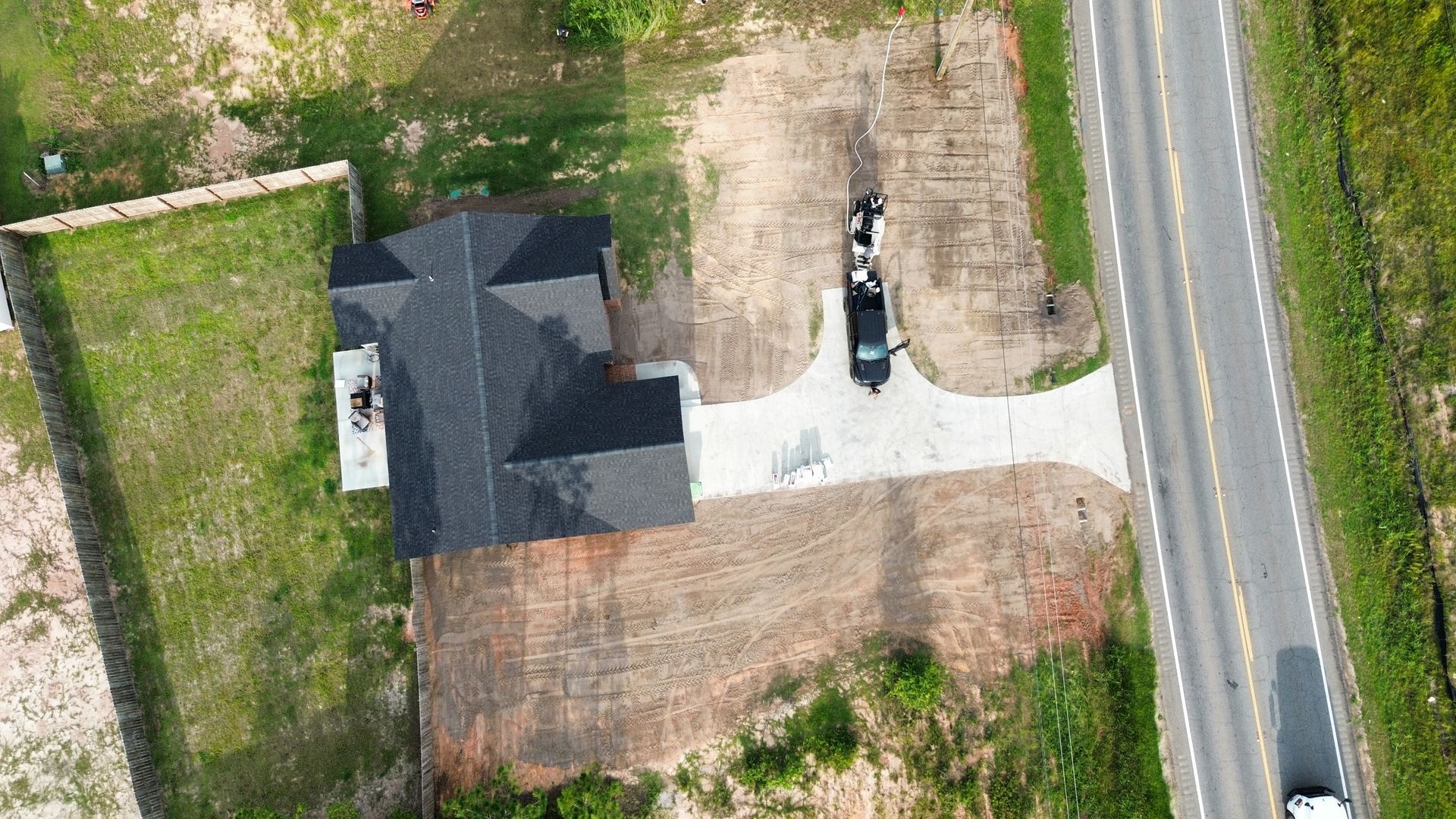 Aerial view of a house with a driveway, parked vehicles, and a road beside grassy land.