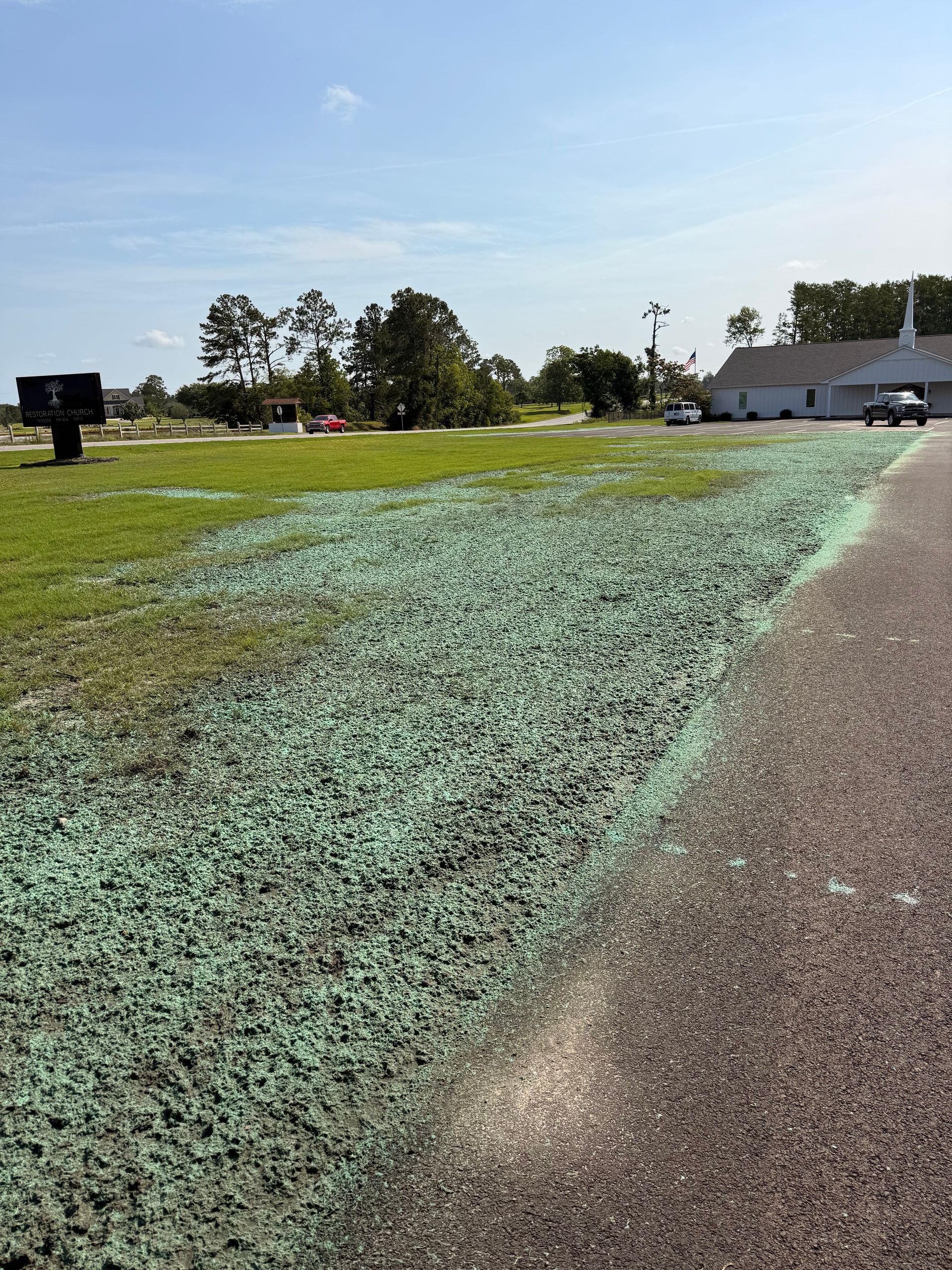 Mowed green field beside a paved path under a clear blue sky, with trees and buildings in the distance