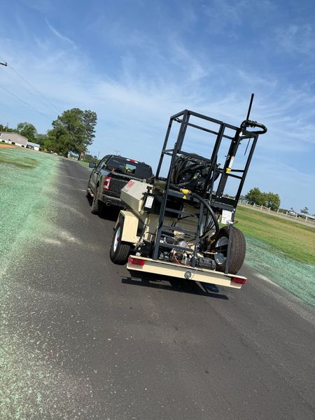 Roadside towing setup with a black utility trailer carrying a machine behind a black car on a sunny rural road.
