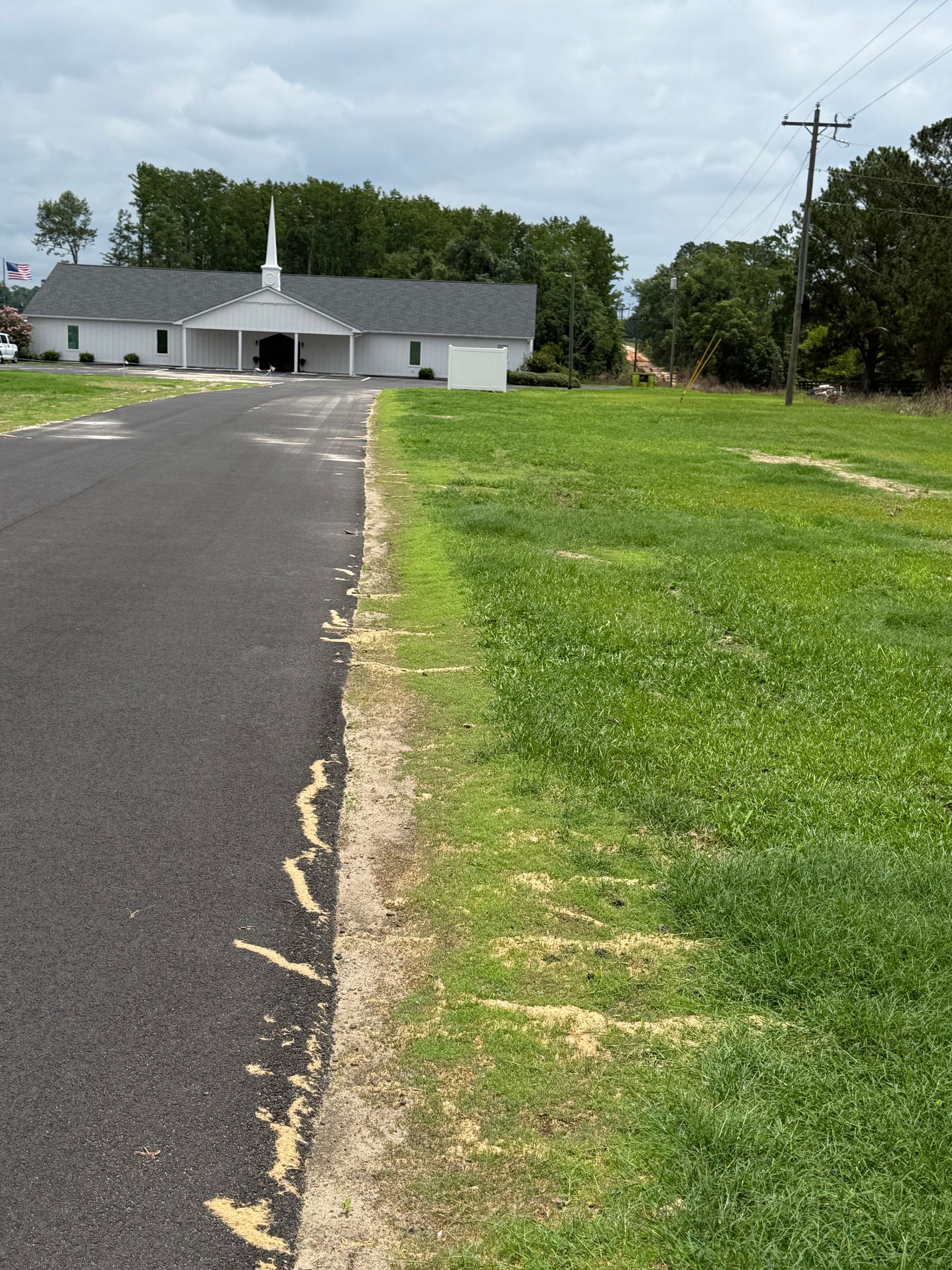 Paved driveway leading to a small white building beside a grassy field under a cloudy sky