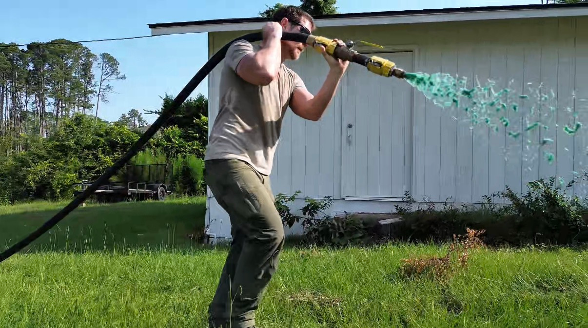 Person outdoors carrying a long pipe on their shoulders while spraying water near a white shed