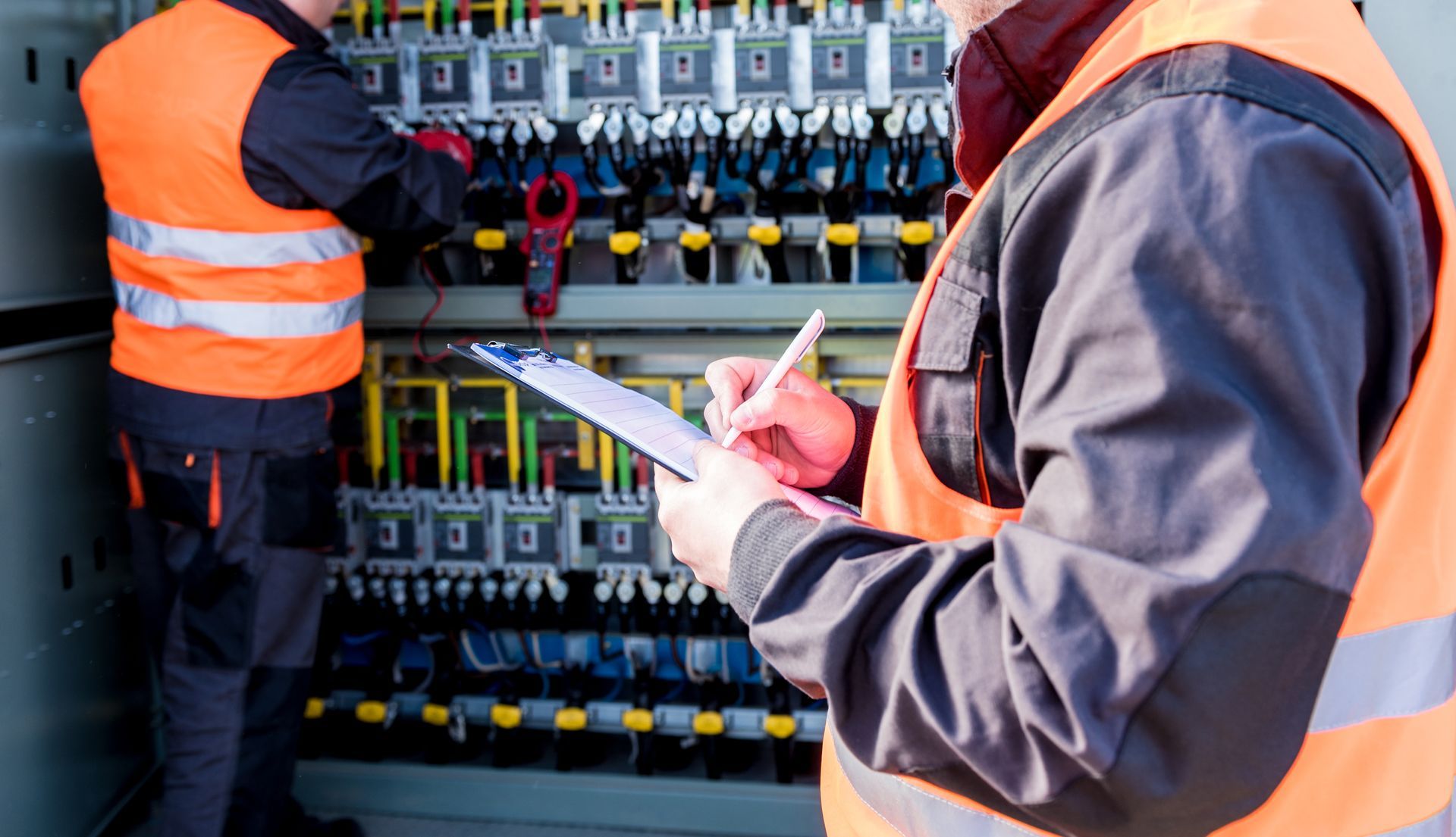 Technicians in orange safety vests inspecting electrical control panels, one taking notes.