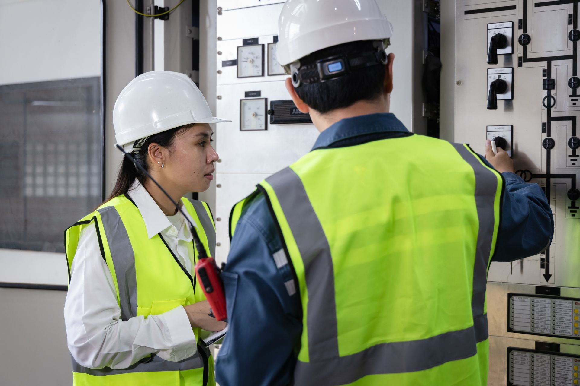 Two workers in hard hats and safety vests inspecting an electrical control panel