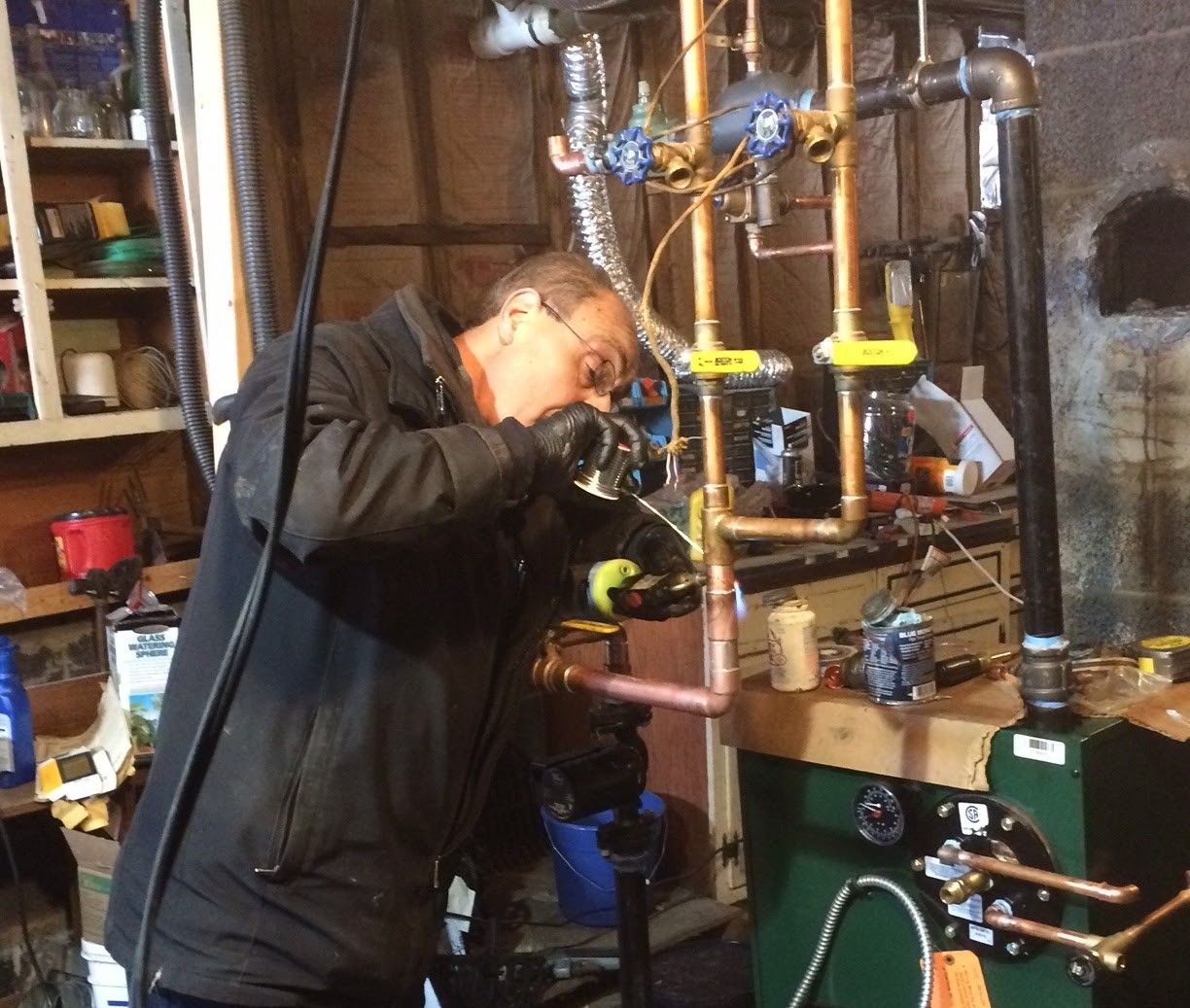 A person wearing gloves works on copper pipes above a green furnace in a basement setting.