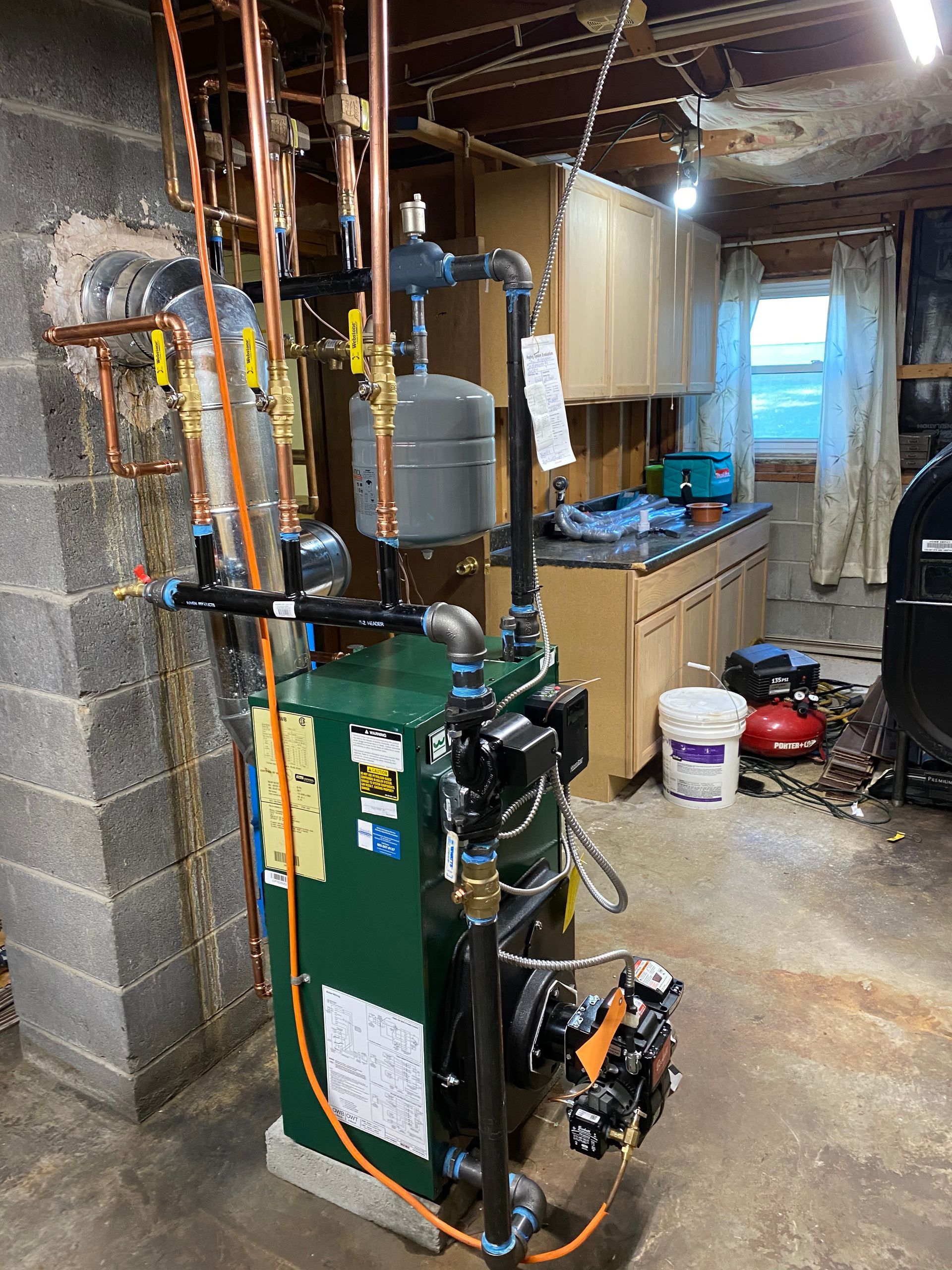Green boiler in a residential basement with copper plumbing, a gray expansion tank, and an oil burner unit.