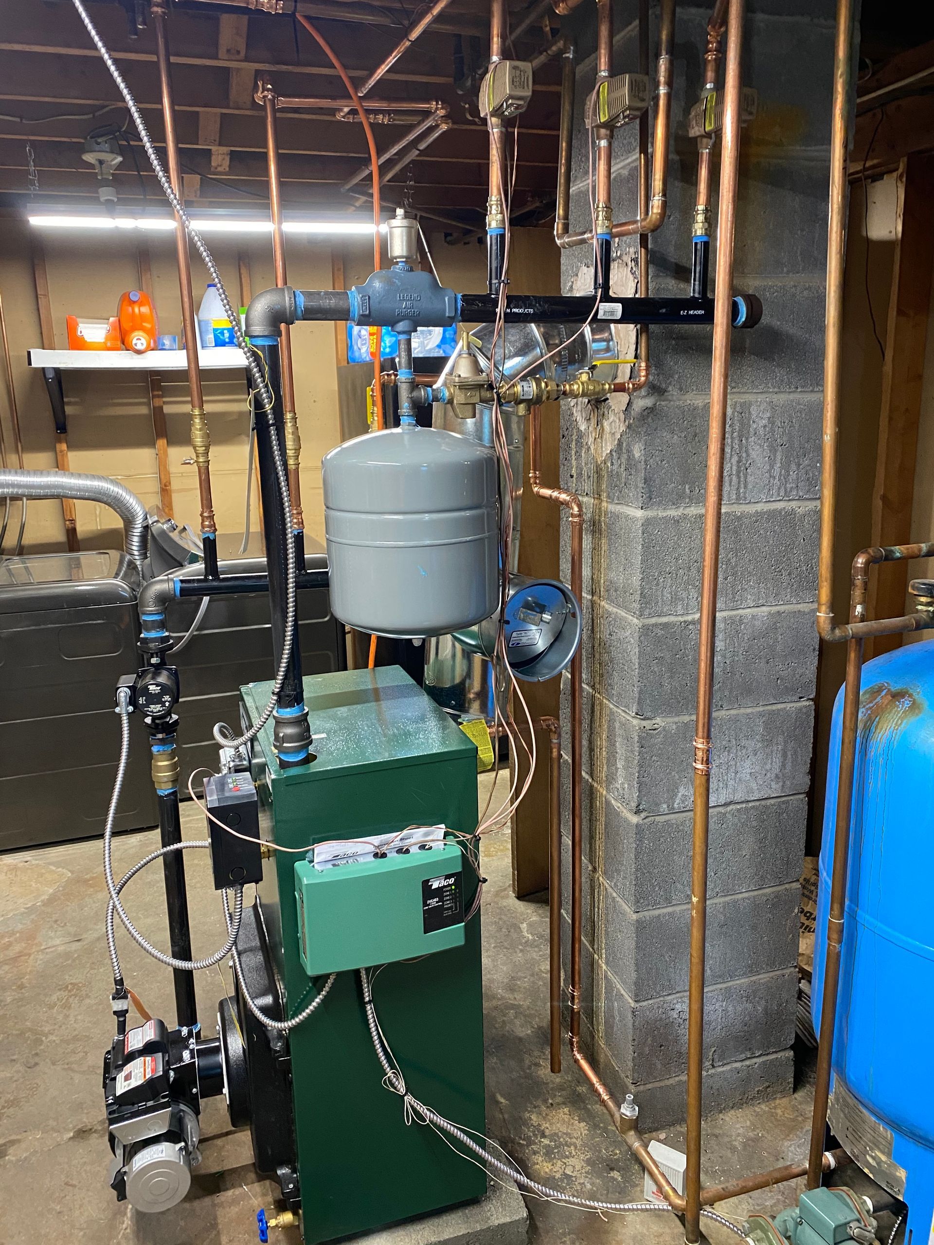 A green residential boiler system with connected copper pipes, a gray expansion tank, and a blue water tank in a basement.