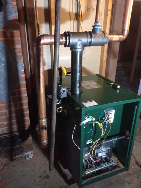 A green residential boiler unit in a basement, connected to overhead copper piping and a central black iron flue.